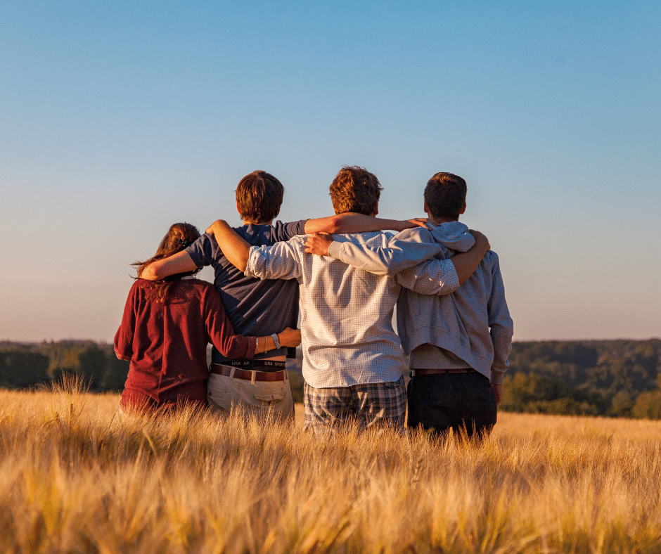 Friends with backs to the camera, looking over a wheat field, holding each other in unity.
