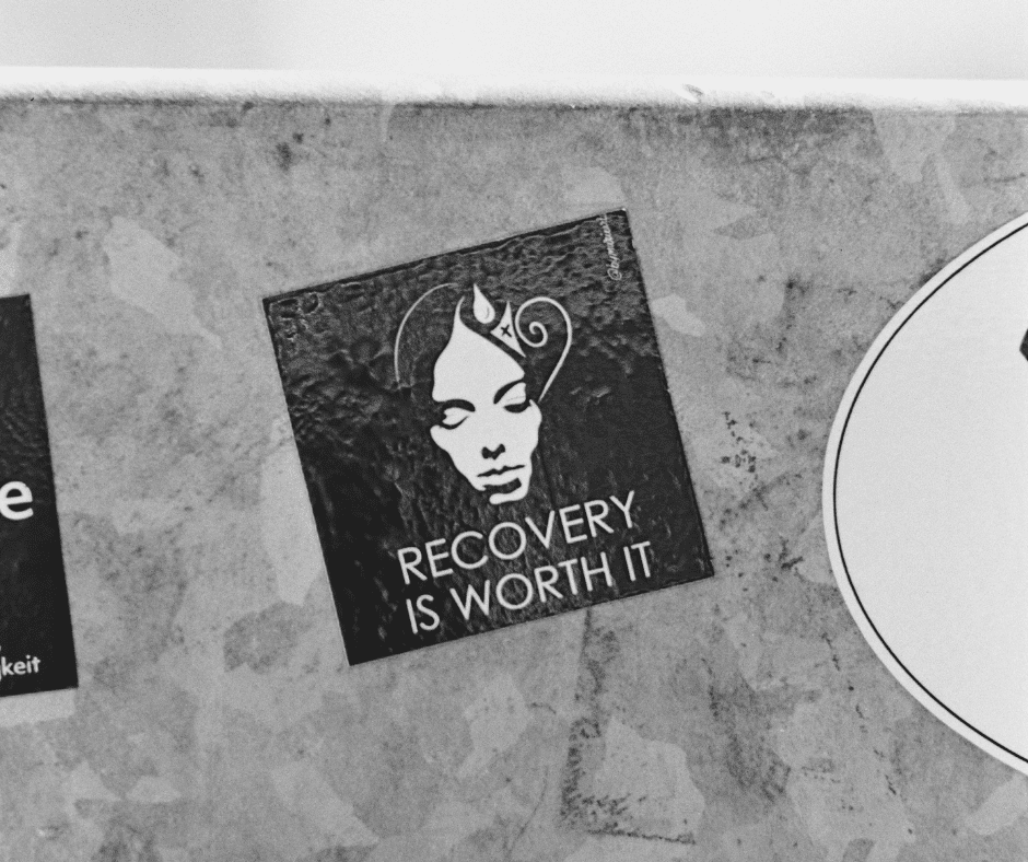 Gray wall with a black sticker showing a face and the text 'Recovery Is Worth It' beneath.
