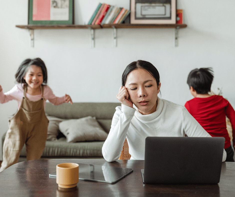 A mother with a bored expression staring down at a laptop screen while her lively children play energetically in the background.