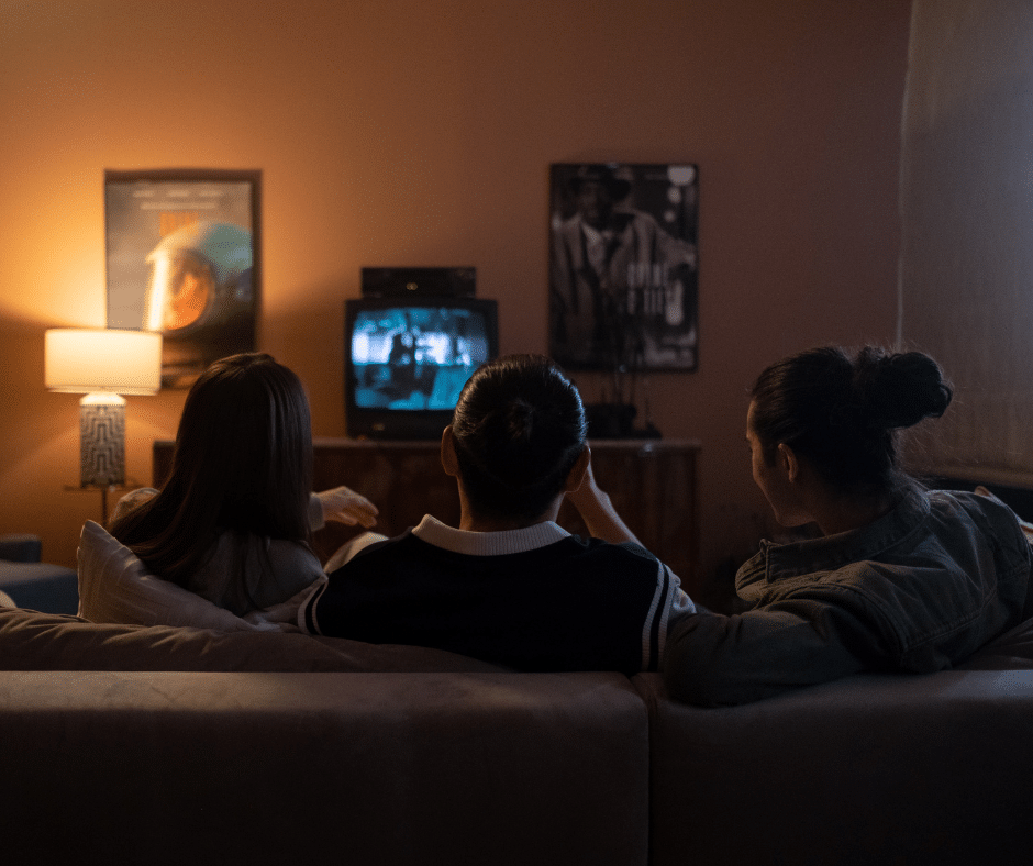 Three friends sitting side-by-side on a couch with their backs turned, focused on watching a television show.