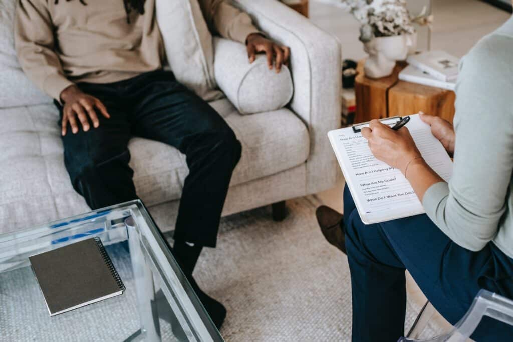 A man in addiction treatment sitting on a couch, engaging in a conversation with a substance use counselor who is attentively taking notes on a clipboard.