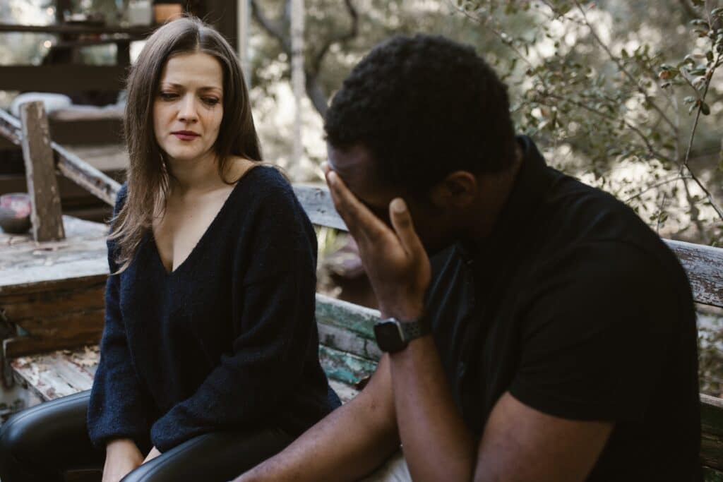 A couple sitting on a park bench, both looking sad, with the woman staring at her husband, reflecting the emotional strain in their relationship.