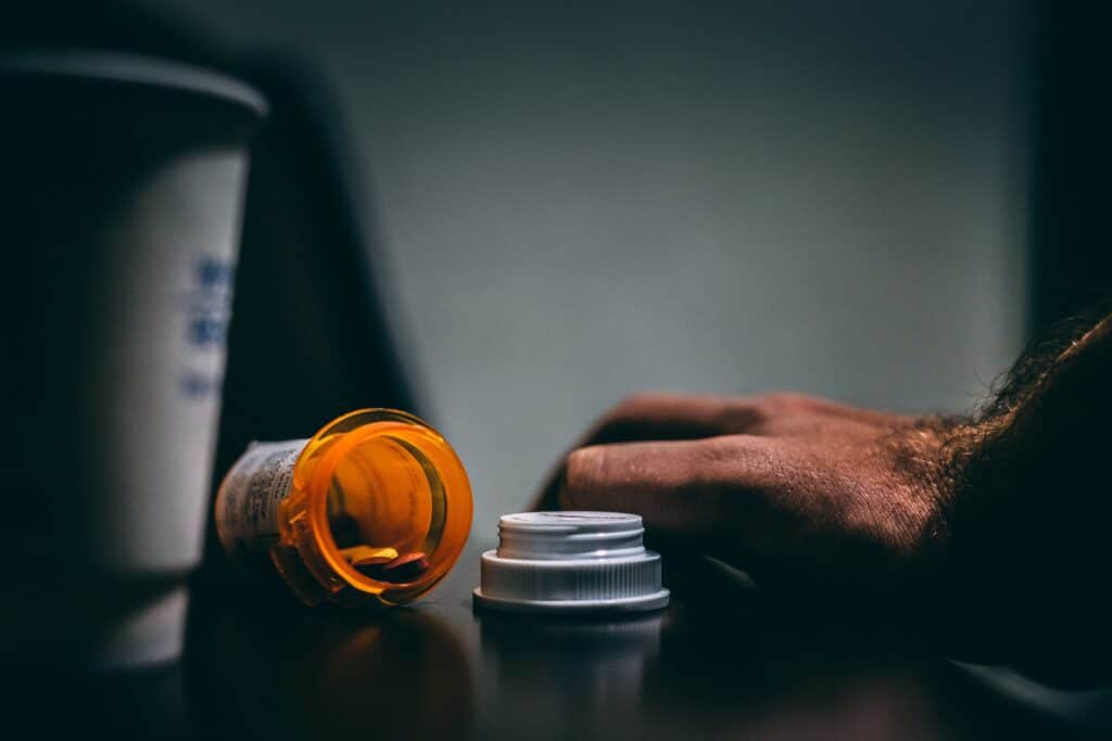 A hand resting on a table beside an open medication bottle, revealing visible pills inside, symbolizing the issue of opioid use, and possibly opioid misuse.