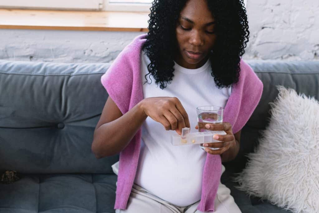 Pregnant mother on a couch, holding a cup of water and a medication pill box, preparing to take FDA-approved buprenorphine (Suboxone), a safe treatment for opioid addiction or opioid use disorder during pregnancy.