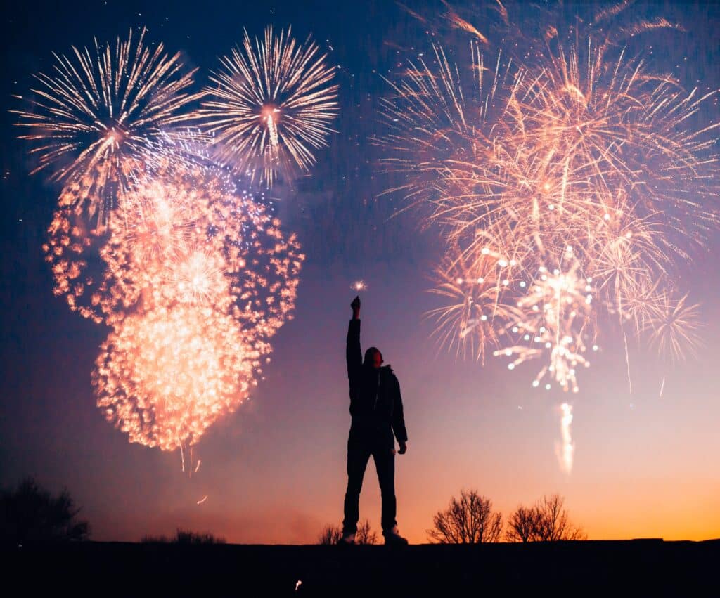 A person holding a sparkler in the air, with vibrant fireworks bursting in the background, symbolizing a joyful commitment to New Year's recovery resolutions.