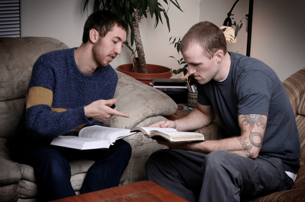 Two men sit on a couch, deeply engaged in a discussion over a book, suggesting a supportive friendship.