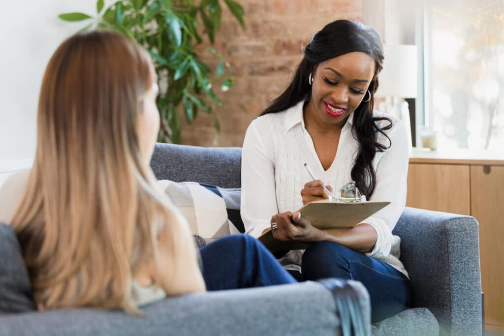 The mid adult female counselor smiles as she takes notes while the unrecognizable young adult woman shares her story.